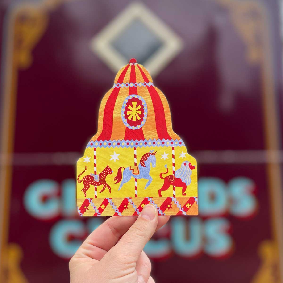 Sustainably made carousel shaped greetings card held by a hand against a Circus wagon