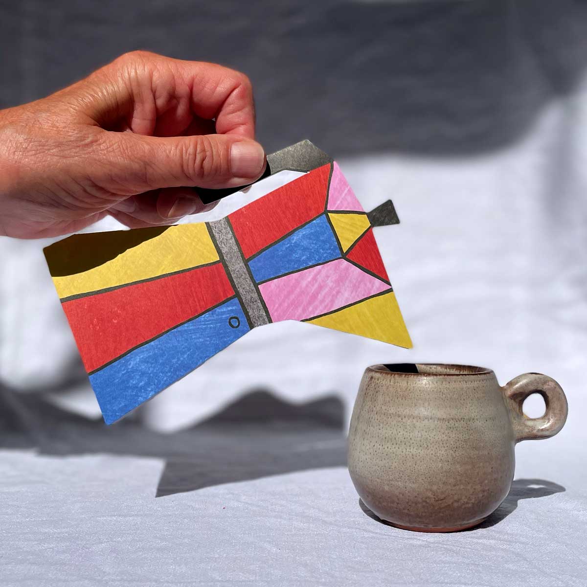 Hand holding a colorful geometric Coffee Pot shaped card next to a ceramic mug on a neutral background