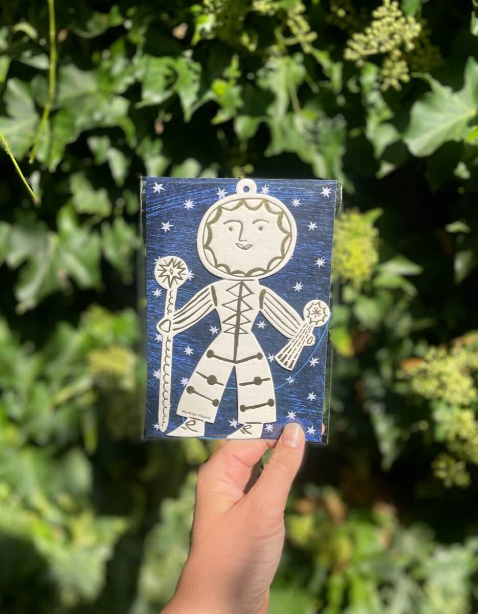 Hand holding a packaged Sun Folk seasonal ornament, surrounded by green foliage.