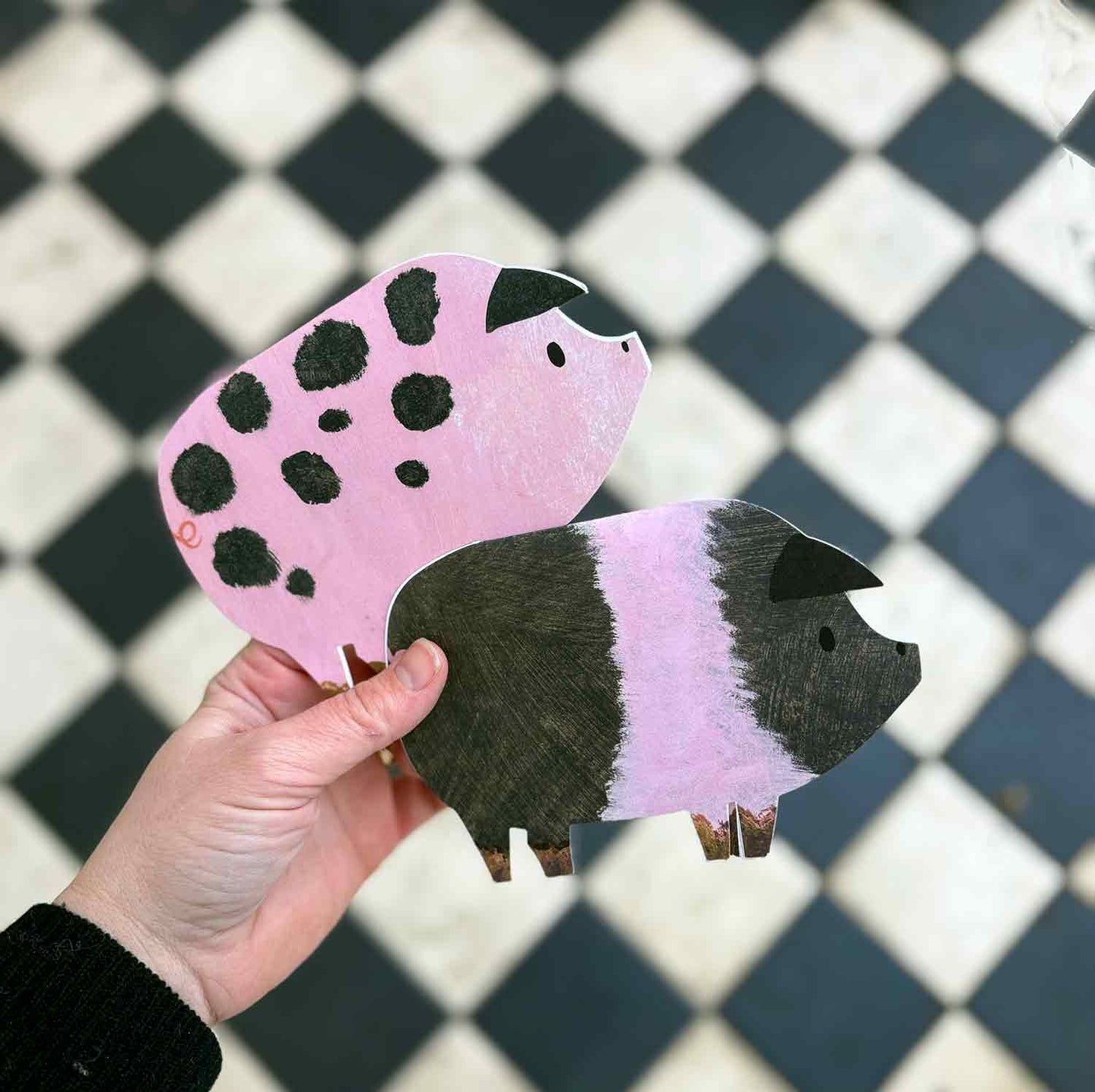 Hand holding a pink and black spotted pig-shaped greetings card and a black and pink striped saddleback pig greetings card against a checkered background