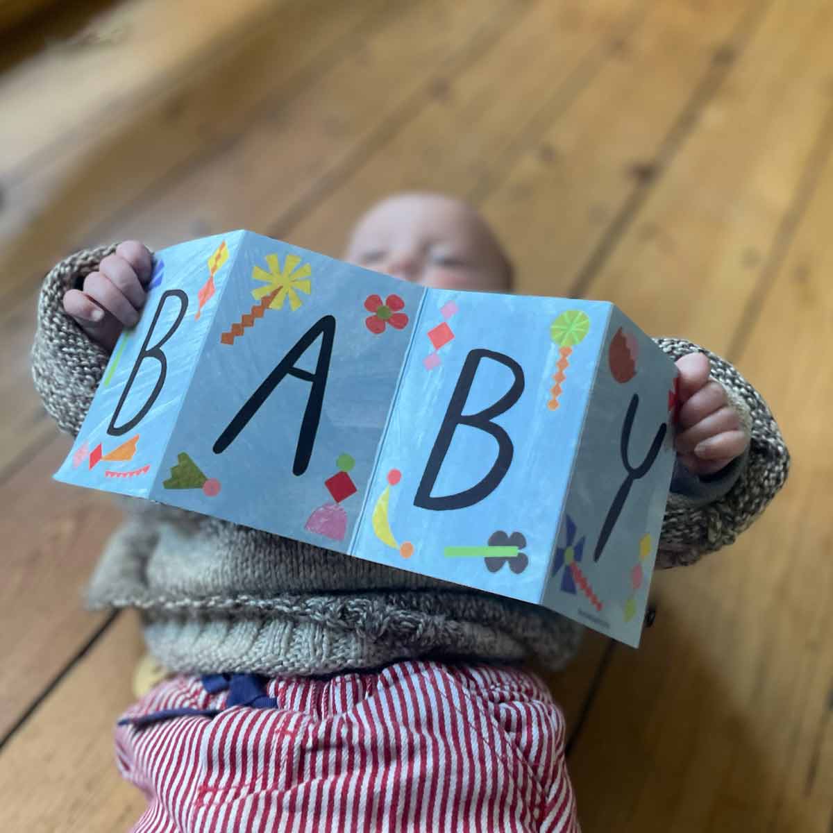 A newborn baby lying down, gently holding a New Baby Concertina Greeting Card. The hand-painted design and eco-friendly recycled paper make it a perfect sustainable card choice for new parents.