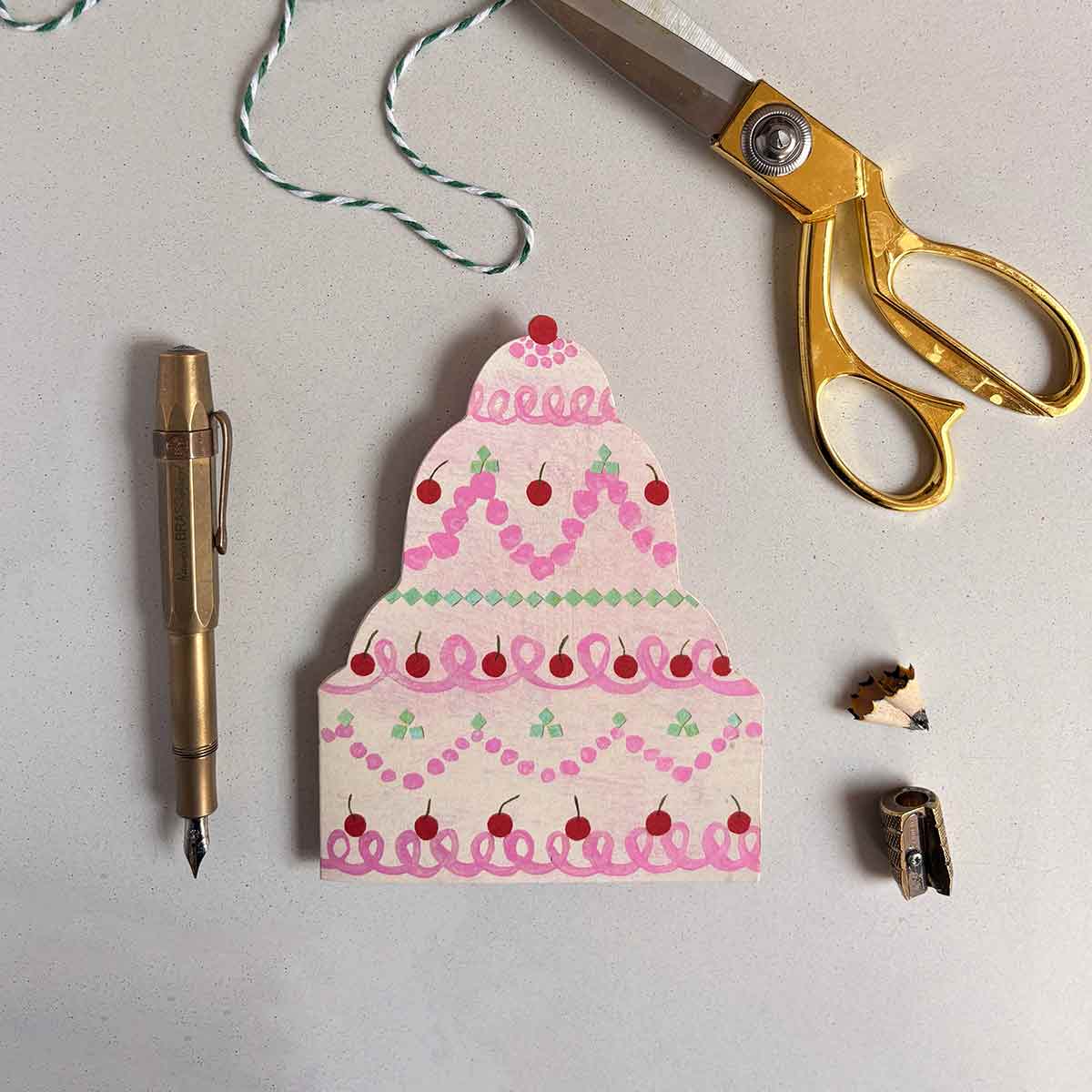 Decorative cake-shaped birthday greetings card  with a pen, scissors, and a pencil sharpener on a gray background.