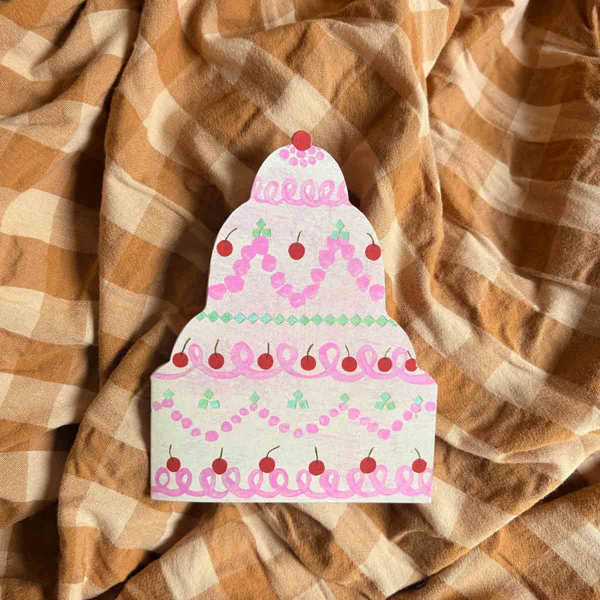 Die cut wedding cake greetings card with pink icing and red cherries on a mustard and ochre checkered fabric background