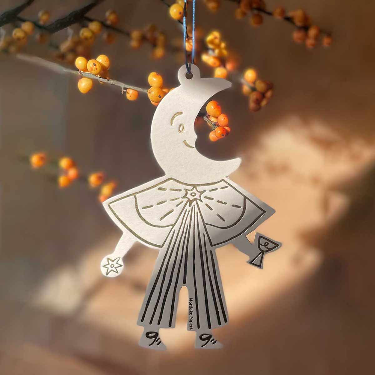Decorative gold and cream card moon shaped christmas ornament hanging from a branch with orange berries.