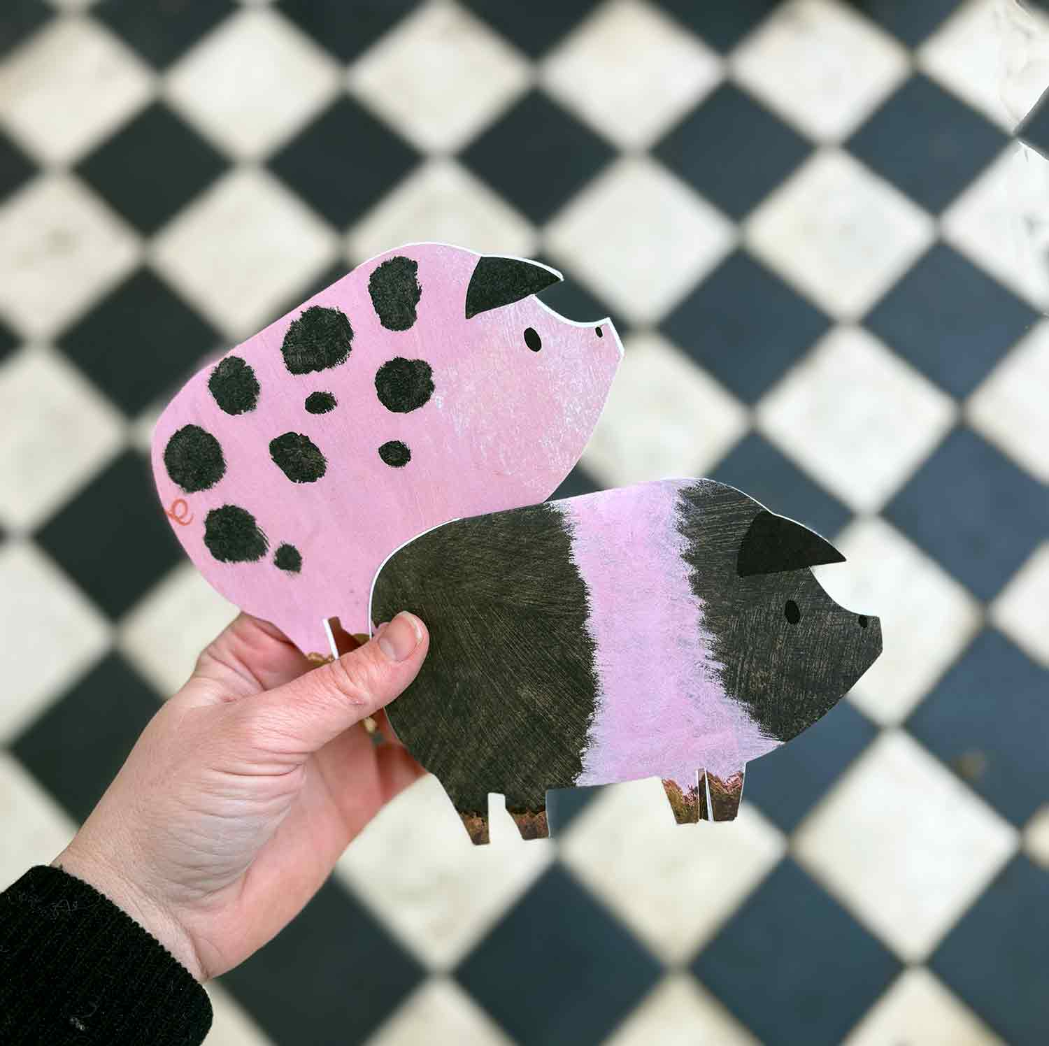 Hand holding a pink and black spotted pig-shaped greetings card and a black and pink striped saddleback pig greetings card against a checkered background