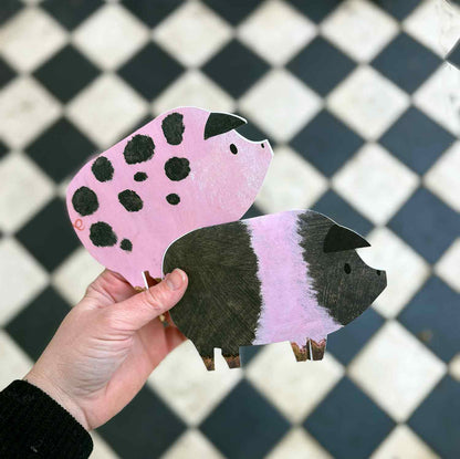 Hand holding a pink and black spotted pig-shaped greetings card and a black and pink striped saddleback pig greetings card against a checkered background