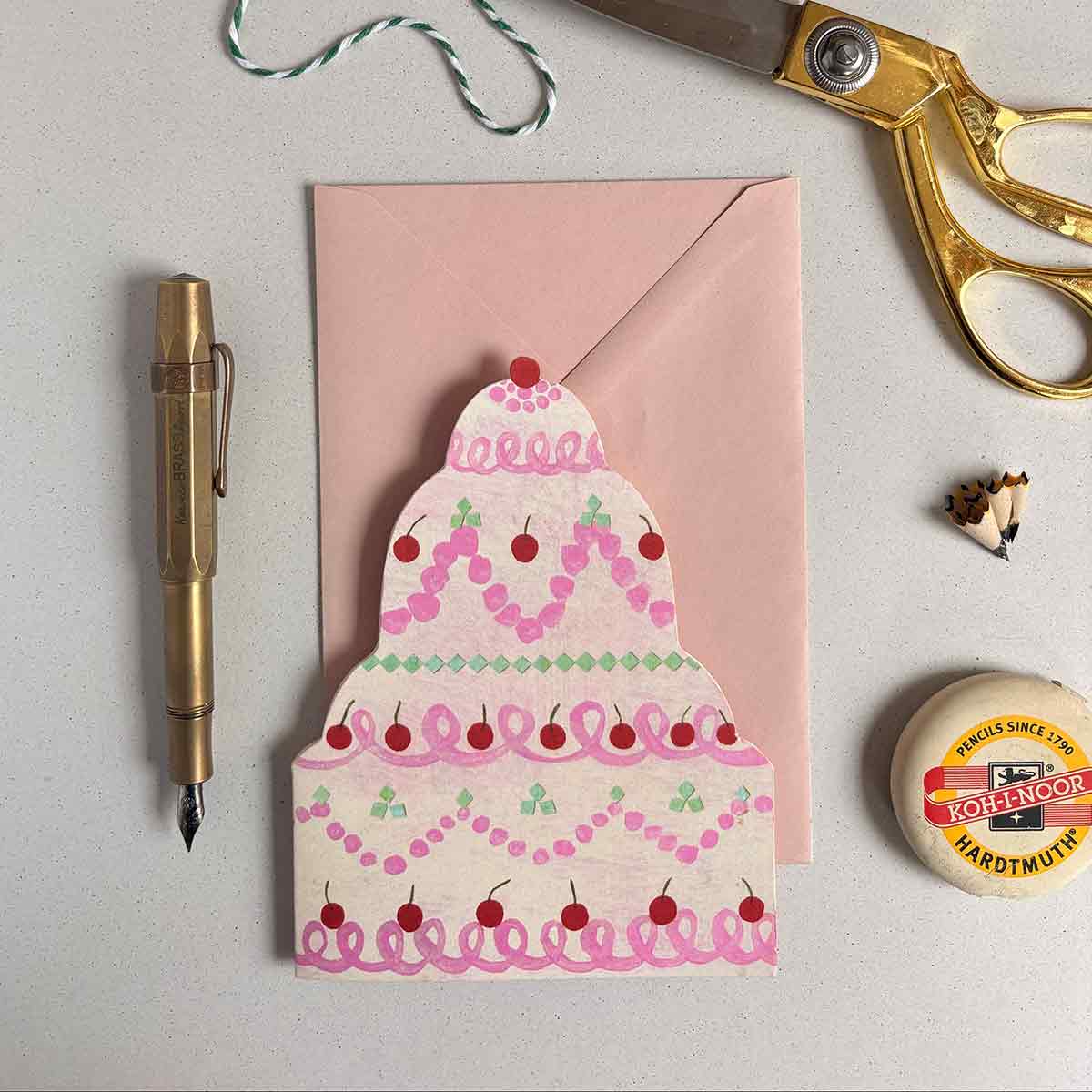 Joyful birthday cake shaped card with pink and pistachio frosting with a pen, scissors, and  an eraser on a light gray background