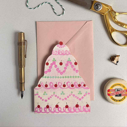 Joyful birthday cake shaped card with pink and pistachio frosting with a pen, scissors, and  an eraser on a light gray background