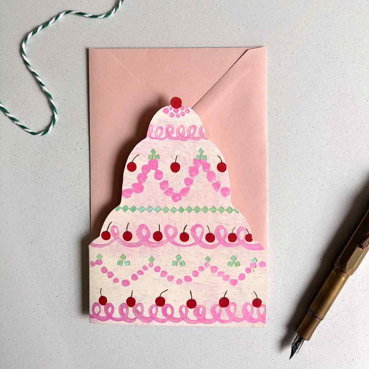 Cream and blush greetings card shaped like a tiered cake with pink icing and red cherries on a light gray background.