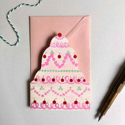 Cream and blush greetings card shaped like a tiered cake with pink icing and red cherries on a light gray background.