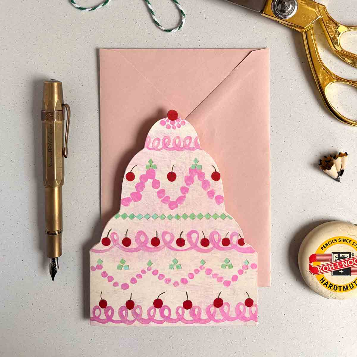 Wedding cake shaped greetings card on a pink envelope, surrounded by a pen, scissors, and an eraser